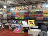A vibrant shop displaying an array of colorful kites and kite strings. The walls are lined with neatly arranged spools of colorful kite strings, organized by color. Various kites with intricate designs, including stars and floral patterns, are displayed prominently on tables and hanging from the ceiling. Two people are seen working in the background, seemingly preparing or selling the kites.