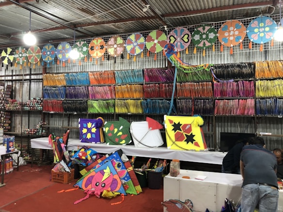 A vibrant shop displaying an array of colorful kites and kite strings. The walls are lined with neatly arranged spools of colorful kite strings, organized by color. Various kites with intricate designs, including stars and floral patterns, are displayed prominently on tables and hanging from the ceiling. Two people are seen working in the background, seemingly preparing or selling the kites.