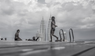 A cloudy sky looms over an infinity pool, where several people in swimwear are relaxing and enjoying the view. In the background, two iconic skyscrapers rise into the sky, providing a striking contrast to the natural environment.