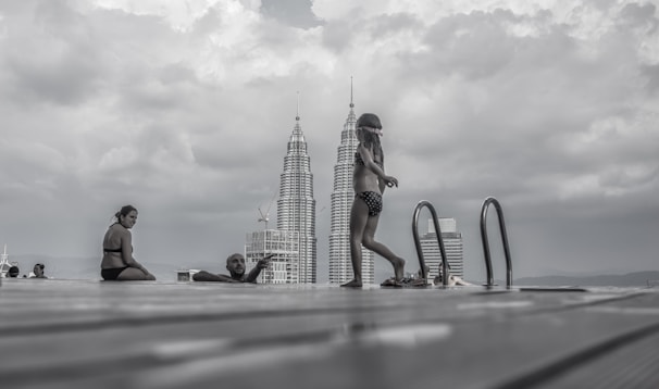 A cloudy sky looms over an infinity pool, where several people in swimwear are relaxing and enjoying the view. In the background, two iconic skyscrapers rise into the sky, providing a striking contrast to the natural environment.