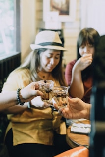 Three people gathered at a table clinking glasses filled with drinks and coffee beans, creating a friendly and intimate atmosphere in a cozy indoor setting. One person is wearing a hat, adding a casual touch to the scene.