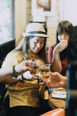 Three people gathered at a table clinking glasses filled with drinks and coffee beans, creating a friendly and intimate atmosphere in a cozy indoor setting. One person is wearing a hat, adding a casual touch to the scene.