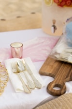 A set of ornate cutlery, including a fork, spoon, and knife, is placed on a white napkin next to a wooden serving board. A pink candle holder is nearby, and in the background, a beverage dispenser with fruit slices is partially visible.
