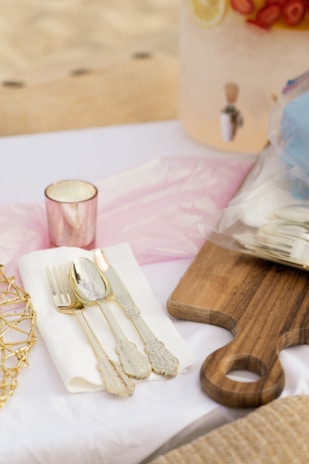 A set of ornate cutlery, including a fork, spoon, and knife, is placed on a white napkin next to a wooden serving board. A pink candle holder is nearby, and in the background, a beverage dispenser with fruit slices is partially visible.