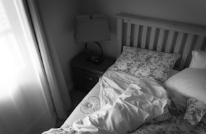 A neatly made bed is positioned near a window with sheer curtains allowing soft light to enter. The bed has a wooden headboard and is covered with floral-patterned pillows. A nightstand is beside the bed with an elegant lamp. The room has a calm and serene appearance.