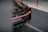 A luxury sports car is partially visible, focusing on the rear end. The car features sleek lines and a glossy black finish with red accents. A license plate and a badge indicating the brand are prominently displayed. The background consists of a road with a section of sidewalk and a barrier.