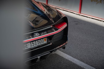 A luxury sports car is partially visible, focusing on the rear end. The car features sleek lines and a glossy black finish with red accents. A license plate and a badge indicating the brand are prominently displayed. The background consists of a road with a section of sidewalk and a barrier.