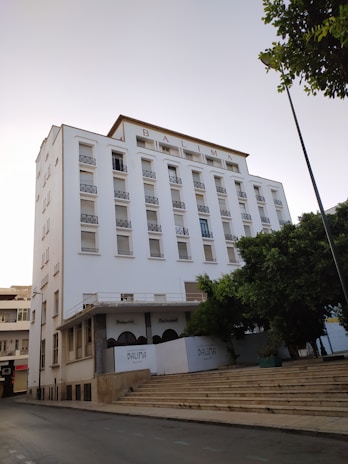 A large multi-story white building with the word 'BALIMA' displayed prominently at the top. It has multiple windows, most of which appear to be closed. There are arched entrances labeled 'Brasserie' and 'Restaurant' on the ground floor. The building is adjacent to a wide set of stone steps and is partially obscured by a tree. The scene seems to be shot during the day with clear skies.