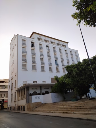 A large multi-story white building with the word 'BALIMA' displayed prominently at the top. It has multiple windows, most of which appear to be closed. There are arched entrances labeled 'Brasserie' and 'Restaurant' on the ground floor. The building is adjacent to a wide set of stone steps and is partially obscured by a tree. The scene seems to be shot during the day with clear skies.