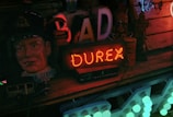 A dimly lit scene depicting a vintage and eclectic collection of objects, including a large red neon sign reading 'Durex'. To the left, there is a mannequin head of a man wearing a cap, and several other items including a canister, letters forming part of a word, and a small vintage toy bus.
