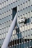 A modern glass building facade with a grid pattern is shown. Two workers are performing maintenance or cleaning on a suspended platform, positioned near a large structural beam that intersects with the building's surface.