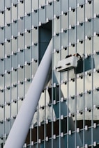 A modern glass building facade with a grid pattern is shown. Two workers are performing maintenance or cleaning on a suspended platform, positioned near a large structural beam that intersects with the building's surface.