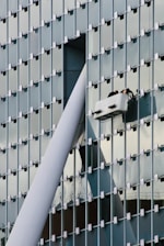A modern glass building facade with a grid pattern is shown. Two workers are performing maintenance or cleaning on a suspended platform, positioned near a large structural beam that intersects with the building's surface.