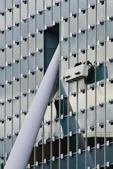 A modern glass building facade with a grid pattern is shown. Two workers are performing maintenance or cleaning on a suspended platform, positioned near a large structural beam that intersects with the building's surface.