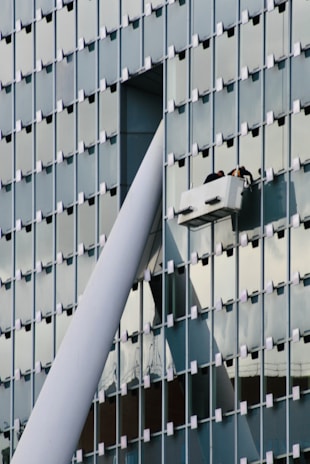 A modern glass building facade with a grid pattern is shown. Two workers are performing maintenance or cleaning on a suspended platform, positioned near a large structural beam that intersects with the building's surface.