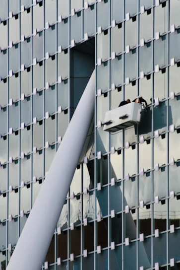 A modern glass building facade with a grid pattern is shown. Two workers are performing maintenance or cleaning on a suspended platform, positioned near a large structural beam that intersects with the building's surface.