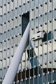 A modern glass building facade with a grid pattern is shown. Two workers are performing maintenance or cleaning on a suspended platform, positioned near a large structural beam that intersects with the building's surface.