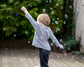 A young child with blonde hair, wearing a striped shirt, joyfully plays with bubbles in an outdoor setting. The child's back is turned to the camera, with one arm raised in excitement amidst the floating bubbles. The background is lush with greenery, suggesting a garden or park environment.