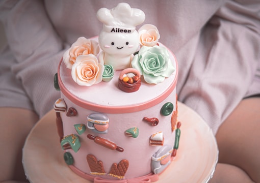 A round, pastel pink cake adorned with fondant decorations including flowers, a small chef figure with a hat labeled 'Aileen,' and various miniature food items like waffle cones and rolling pins. The cake sits on a lightly colored plate, with additional fondant details around the sides.
