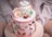 A round, pastel pink cake adorned with fondant decorations including flowers, a small chef figure with a hat labeled 'Aileen,' and various miniature food items like waffle cones and rolling pins. The cake sits on a lightly colored plate, with additional fondant details around the sides.