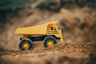 A yellow toy dump truck positioned on a mound of dirt, with its wheels partially covered in soil. The truck has a bold black stripe and text along its side. The background consists of a blurred earthy surface, suggesting an outdoor, construction-like setting.