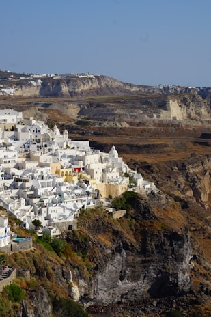 Whitewashed buildings with blue domes perched on a rugged cliff overlooking the sea, characteristic of a Greek island setting. The landscape features dramatic cliffs and a winding coastline, with sparse vegetation and earthy tones in the background.