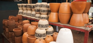 Various clay pots are displayed on a red metal table. Some pots are plain terracotta, while others feature intricate black and white patterns. The arrangement includes different shapes and sizes, suggesting they are for sale at a market or craft fair. A set of tools can be seen on the right.