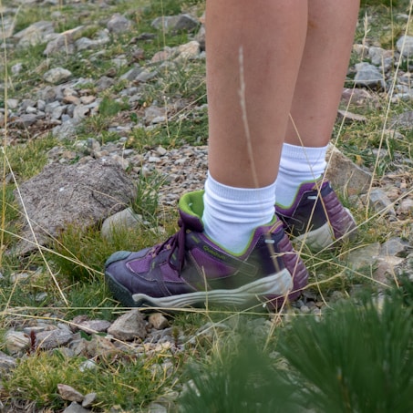 A person is standing outdoors on rocky terrain, wearing white socks and purple hiking shoes with green accents. The ground is covered with small rocks and sparse patches of grass, suggesting a natural setting such as a trail or hillside.