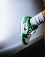 A green and white sneaker with a black swoosh logo is worn on a foot. The foot is against a reflective white surface, and the sock is white with a black detail.