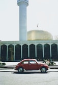 A red vintage car is parked in front of an Islamic building with a prominent golden dome and a tall minaret. The architecture features a series of arched windows, and there's a crescent moon symbol atop the dome. The scene is bright and the sky is clear.