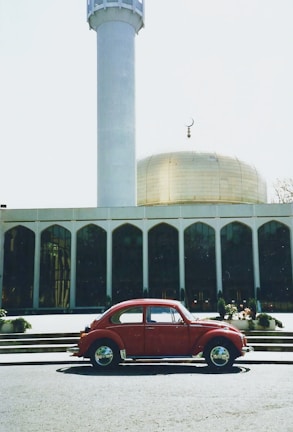 A red vintage car is parked in front of an Islamic building with a prominent golden dome and a tall minaret. The architecture features a series of arched windows, and there's a crescent moon symbol atop the dome. The scene is bright and the sky is clear.