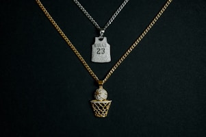 Two chains with pendants are displayed against a dark background. One chain is gold with a pendant in the shape of a basketball going into a net, embellished with diamonds. The other chain is silver and features a pendant shaped like a basketball jersey with the number '23' and 'BULLS' inscribed on it, also adorned with diamonds.