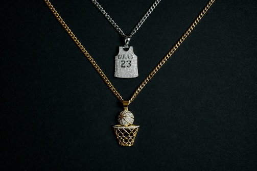 Two chains with pendants are displayed against a dark background. One chain is gold with a pendant in the shape of a basketball going into a net, embellished with diamonds. The other chain is silver and features a pendant shaped like a basketball jersey with the number '23' and 'BULLS' inscribed on it, also adorned with diamonds.