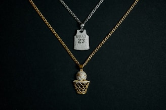 Two chains with pendants are displayed against a dark background. One chain is gold with a pendant in the shape of a basketball going into a net, embellished with diamonds. The other chain is silver and features a pendant shaped like a basketball jersey with the number '23' and 'BULLS' inscribed on it, also adorned with diamonds.