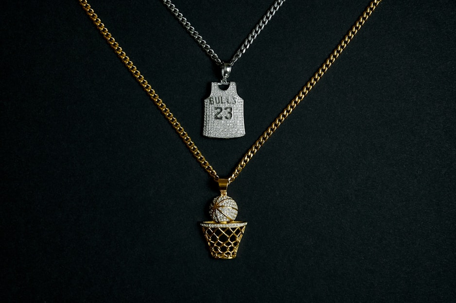 Two chains with pendants are displayed against a dark background. One chain is gold with a pendant in the shape of a basketball going into a net, embellished with diamonds. The other chain is silver and features a pendant shaped like a basketball jersey with the number '23' and 'BULLS' inscribed on it, also adorned with diamonds.