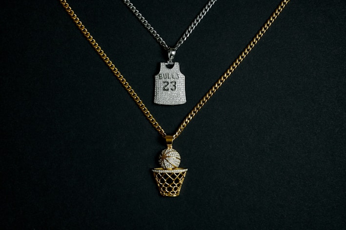 Two chains with pendants are displayed against a dark background. One chain is gold with a pendant in the shape of a basketball going into a net, embellished with diamonds. The other chain is silver and features a pendant shaped like a basketball jersey with the number '23' and 'BULLS' inscribed on it, also adorned with diamonds.