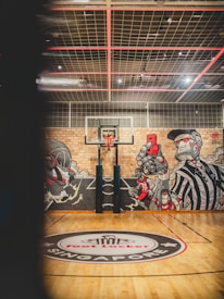 A basketball court featuring a central red and white emblem on the wooden floor, marked with the words 'Foot Locker Singapore'. The brick wall in the background displays vibrant graffiti art with stylized figures, including a basketball player and a referee. Overhead, the ceiling grid is visible with spotlights.