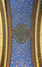 A decorative ceiling panel adorned with intricate floral and geometric patterns. The central design is a complex geometric shape with detailed gold patterns, surrounded by a blue field featuring stylized golden flowers and vines. The panel is bordered by ornate red and gold designs, with green and yellow elements on each side.