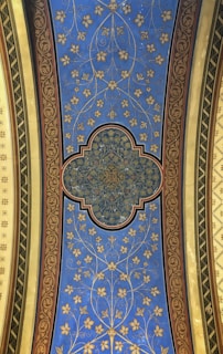 A decorative ceiling panel adorned with intricate floral and geometric patterns. The central design is a complex geometric shape with detailed gold patterns, surrounded by a blue field featuring stylized golden flowers and vines. The panel is bordered by ornate red and gold designs, with green and yellow elements on each side.