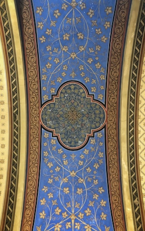 A decorative ceiling panel adorned with intricate floral and geometric patterns. The central design is a complex geometric shape with detailed gold patterns, surrounded by a blue field featuring stylized golden flowers and vines. The panel is bordered by ornate red and gold designs, with green and yellow elements on each side.