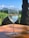 A glass of white wine sits on a wooden table, with condensation visible on the glass. In the background, there is a lush vineyard and a mountainous landscape under a clear blue sky.