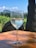 A glass of white wine sits on a wooden table, with condensation visible on the glass. In the background, there is a lush vineyard and a mountainous landscape under a clear blue sky.