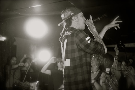 A tattooed individual holding a microphone performs on stage in front of an enthusiastic audience. Behind him, a bright light shines, highlighting the energy of the scene. The person is wearing a checkered shirt and a backward cap, and various audience members can be seen taking photos with smartphones.