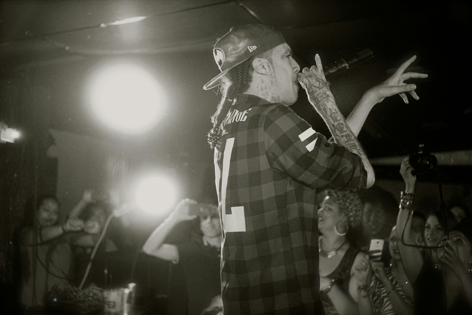 A tattooed individual holding a microphone performs on stage in front of an enthusiastic audience. Behind him, a bright light shines, highlighting the energy of the scene. The person is wearing a checkered shirt and a backward cap, and various audience members can be seen taking photos with smartphones.