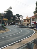 A winding road curves smoothly to the right, bordered by metal guardrails and dry grass. In the background, colorful flags decorate the roadside near a vibrant building with domed architecture. Trees with dense foliage provide a backdrop, and several signs are visible, including one with the name Garuda.