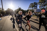 A group of runners participate in a race on a sunny day, with the sun low in the sky casting long shadows. One runner in the foreground is adjusting her gear and another interacts with a race official who is handing out cups. There are spectators and other runners in the background alongside tree-lined streets and distant buildings.