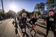 A group of runners participate in a race on a sunny day, with the sun low in the sky casting long shadows. One runner in the foreground is adjusting her gear and another interacts with a race official who is handing out cups. There are spectators and other runners in the background alongside tree-lined streets and distant buildings.