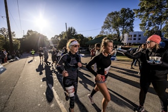 A group of runners participate in a race on a sunny day, with the sun low in the sky casting long shadows. One runner in the foreground is adjusting her gear and another interacts with a race official who is handing out cups. There are spectators and other runners in the background alongside tree-lined streets and distant buildings.