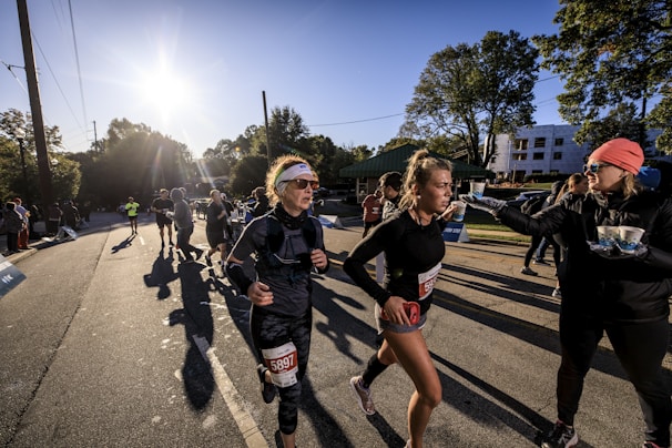 A group of runners participate in a race on a sunny day, with the sun low in the sky casting long shadows. One runner in the foreground is adjusting her gear and another interacts with a race official who is handing out cups. There are spectators and other runners in the background alongside tree-lined streets and distant buildings.