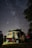 A vintage camper van is parked under a sky full of stars, with the Milky Way clearly visible. The van is illuminated softly, suggesting it is nighttime, and is surrounded by other vehicles and trees. Inside, the van appears to be lived in, with clothes and other belongings visible.